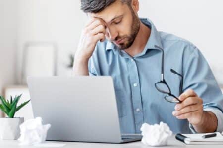 Man working at desk in pain. He is wearing blue shirt and holding glasses. Osteopathoften treat neck pain.