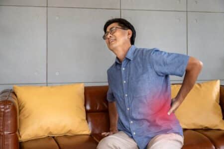 Man wearing blue shirt sitting on yellow couch holding lower back in pain