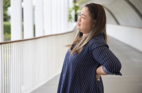 Lady with lower back pain, bulging discs. She is wearing a dark blue top and in pain. she is clutching her lower back on the left side due to sharp pain.