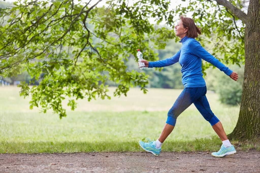 Elderly woman wearing blue work out gear walking outdoors in green park feeling strong and healthy