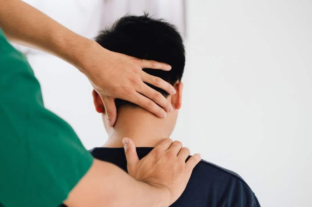 Young male patient sittign on a treatment table with osteopath standing behind him. The osteo is feeling his neck to establish pain or tightness. Patient is wearing a black T shirt.