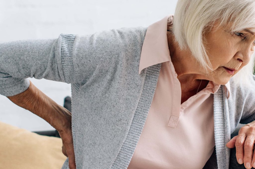An elderly lady with pain from arthritis. She is wearing a grey sweater. Arthritis pain decreases movement. She looks stiff and in pain. osteoarthritis lumbar pain