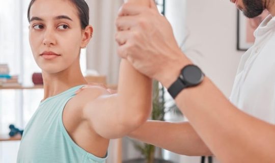 female patient sitting on table with doctor holding left arm up to stretch rotator cuff. Testing for rotator cuff injury. Lady is wearing a light green singlet. Rotator cuff injury, shoulder bursitis, shoulder pain test.