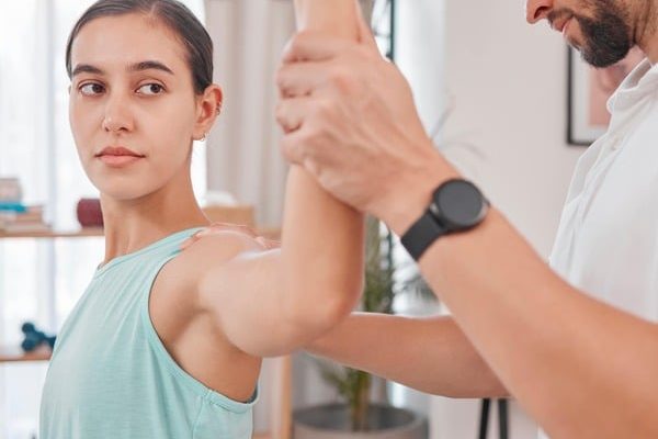 female patient sitting on table with doctor holding left arm up to stretch rotator cuff. Testing for rotator cuff injury. Lady is wearing a light green singlet. Rotator cuff injury, shoulder bursitis, shoulder pain test.