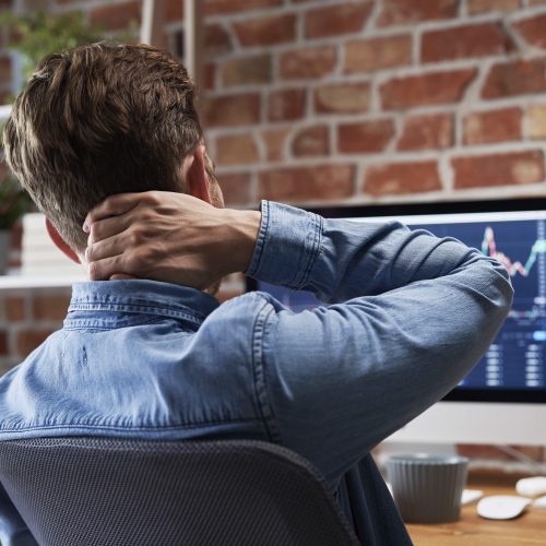 A young man sits at a desk holding his neck in pain. He has compute screens in front of him. He is wearing a blue shirt. neck pain and headaches are represented by hand position.